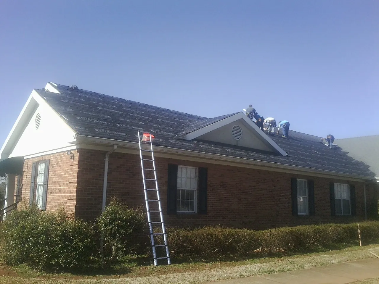 Crew tearing shingles off a brick ranch roof with a ladder set — Metal Roof Repair work by our crew in Zebulon