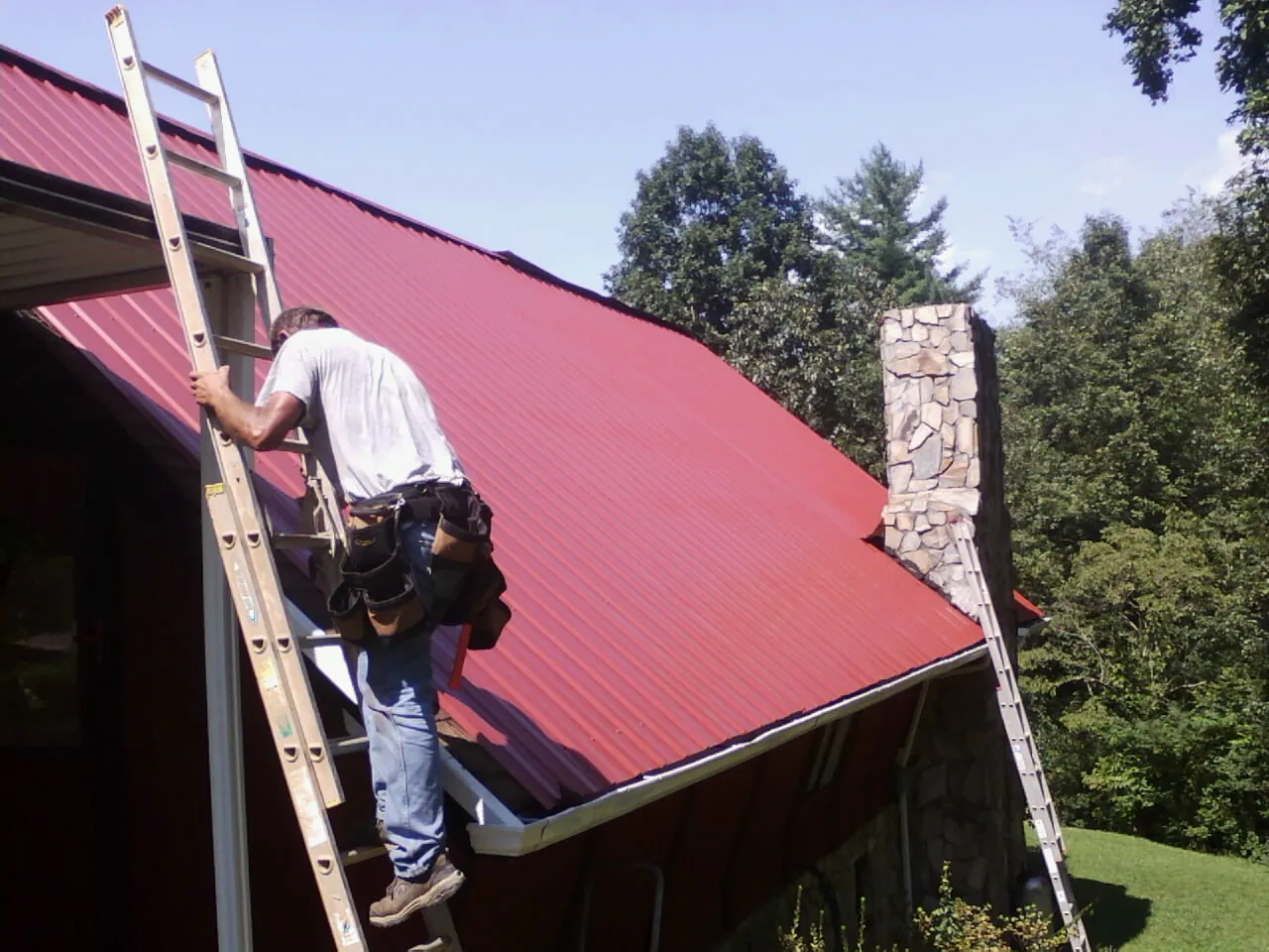 Roofer on a ladder beside a red metal roof and stone chimney — Soffit & Fascia Repair by Roofing Brothers in Zebulon